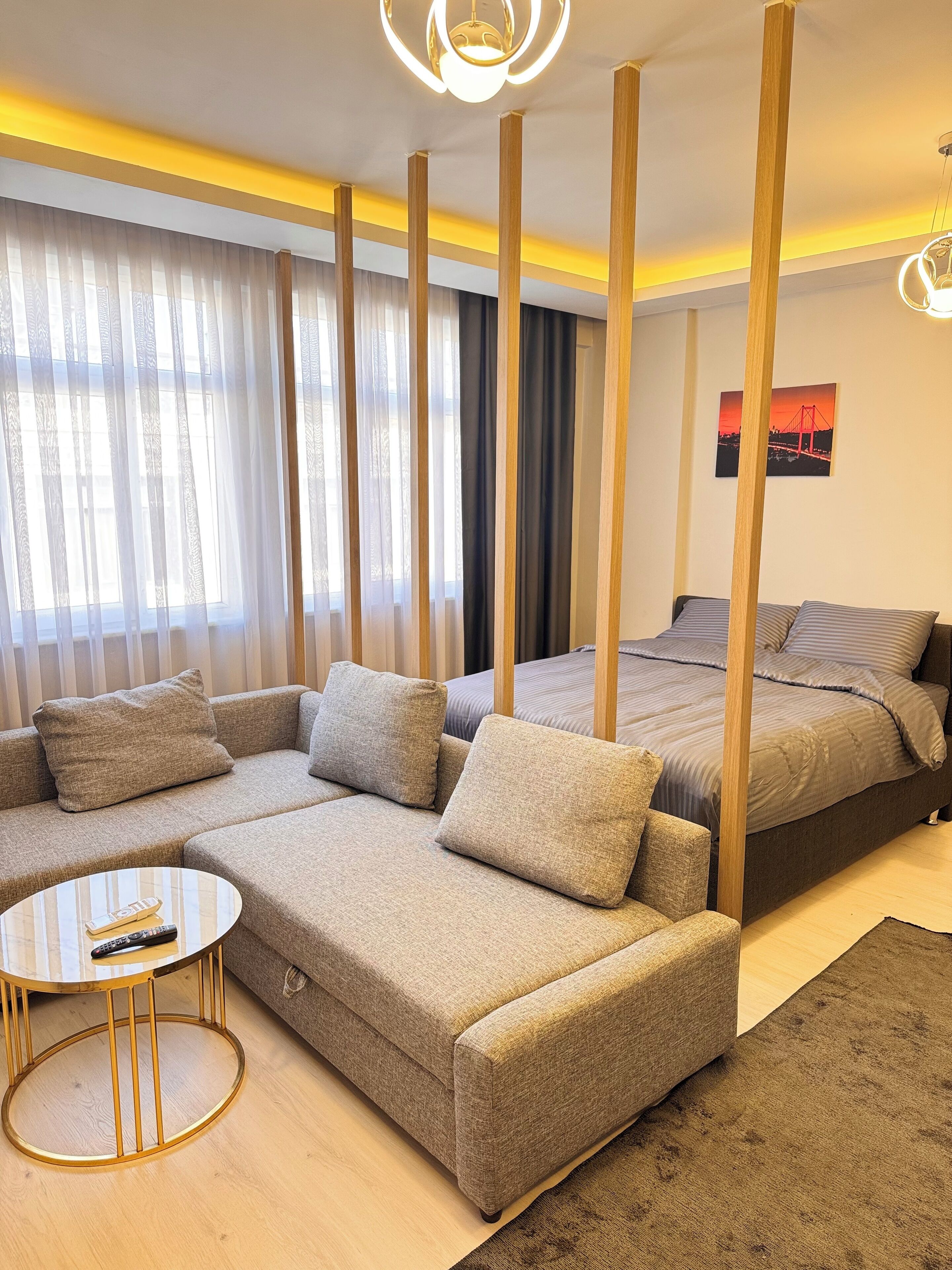 Quarto moderno com uma cama grande, um sofá, uma mesa de centro redonda e uma janela ampla com cortinas leves.