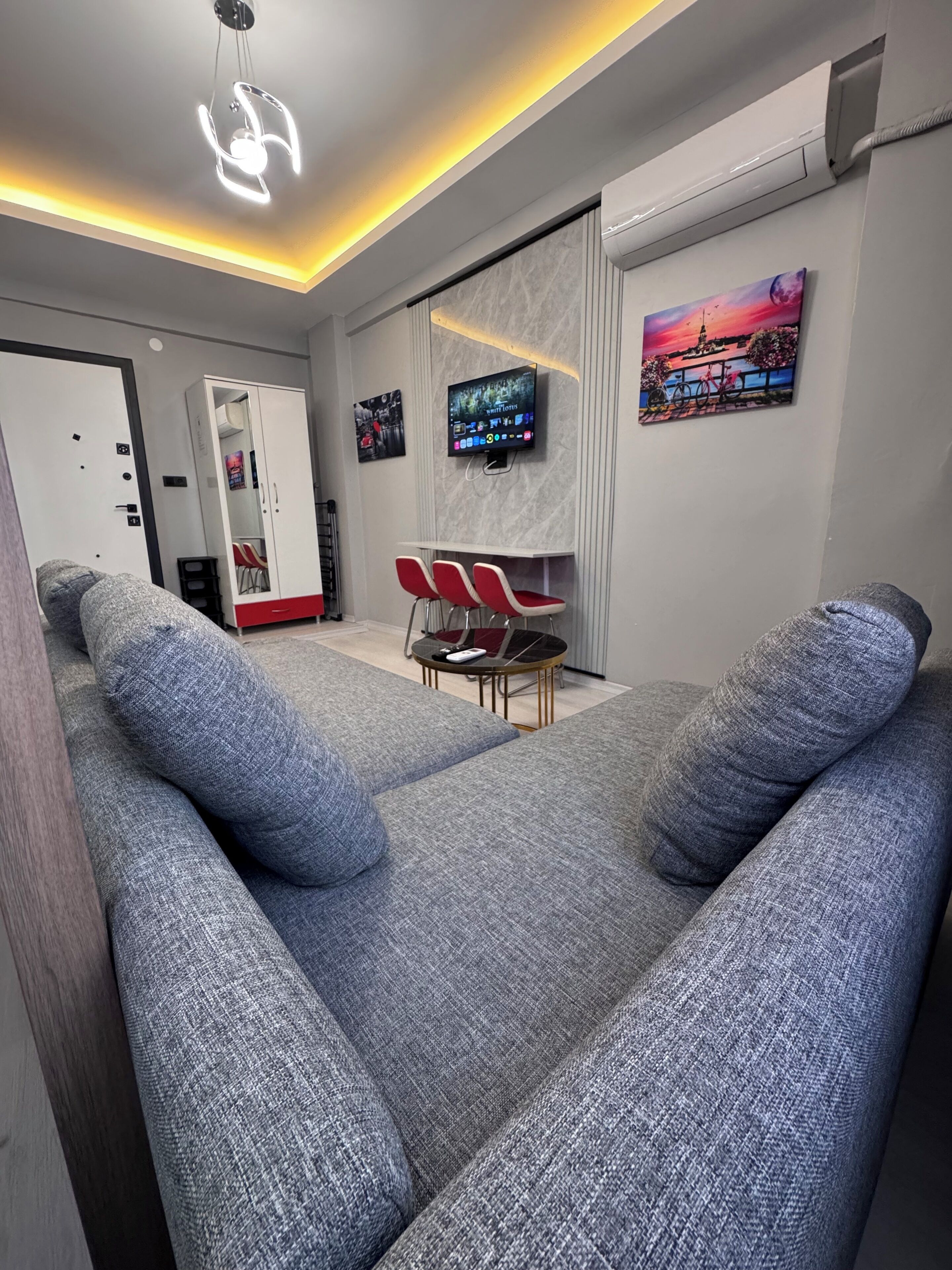 A modern living room with grey sofas, a flat-screen TV mounted on the wall, and a small dining table with red chairs.
