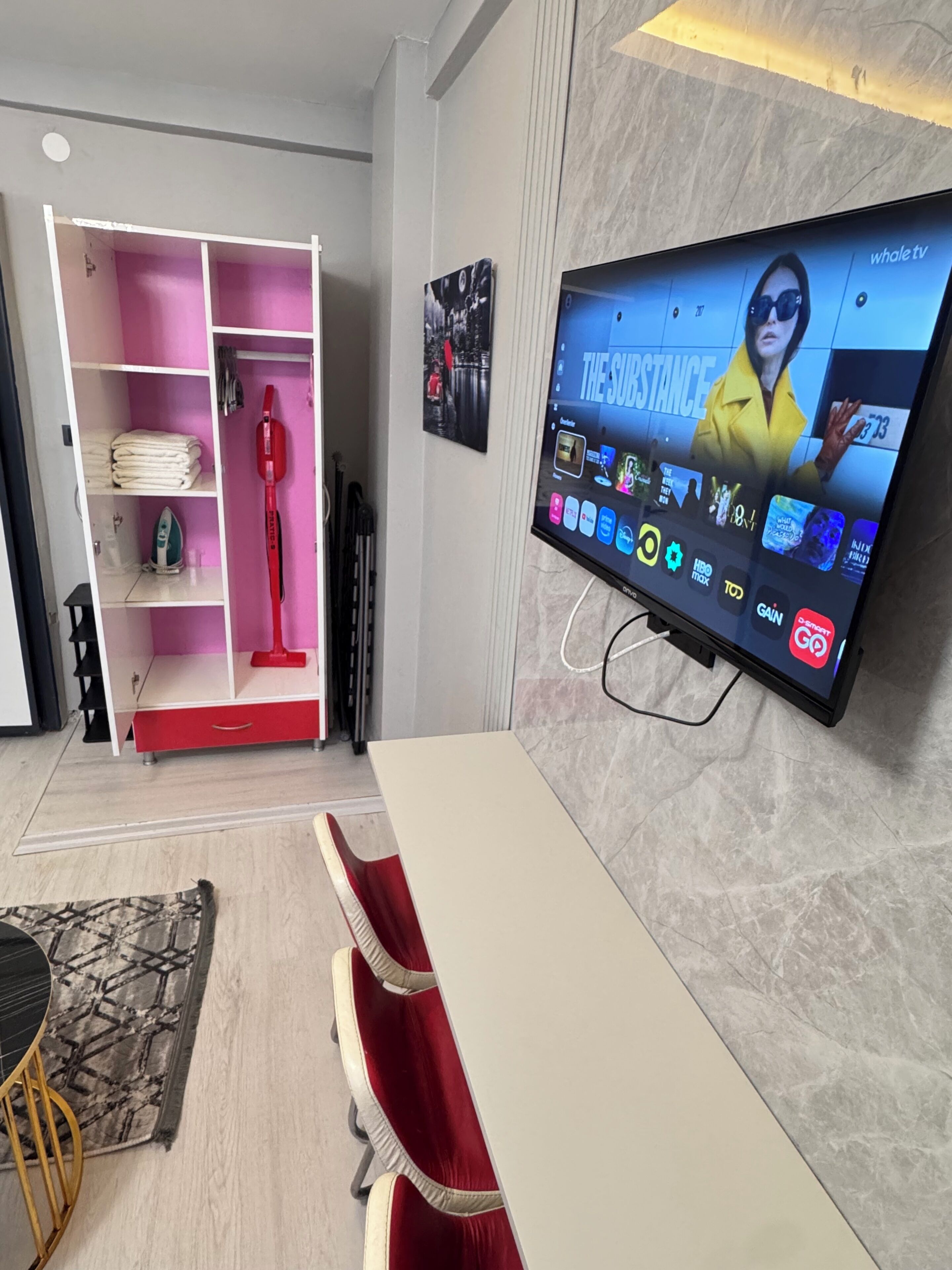 Una sala moderna con un televisor de pantalla plana montado en la pared, un mueble de entretenimiento rojo y blanco, y un comedor con mesa y sillas.