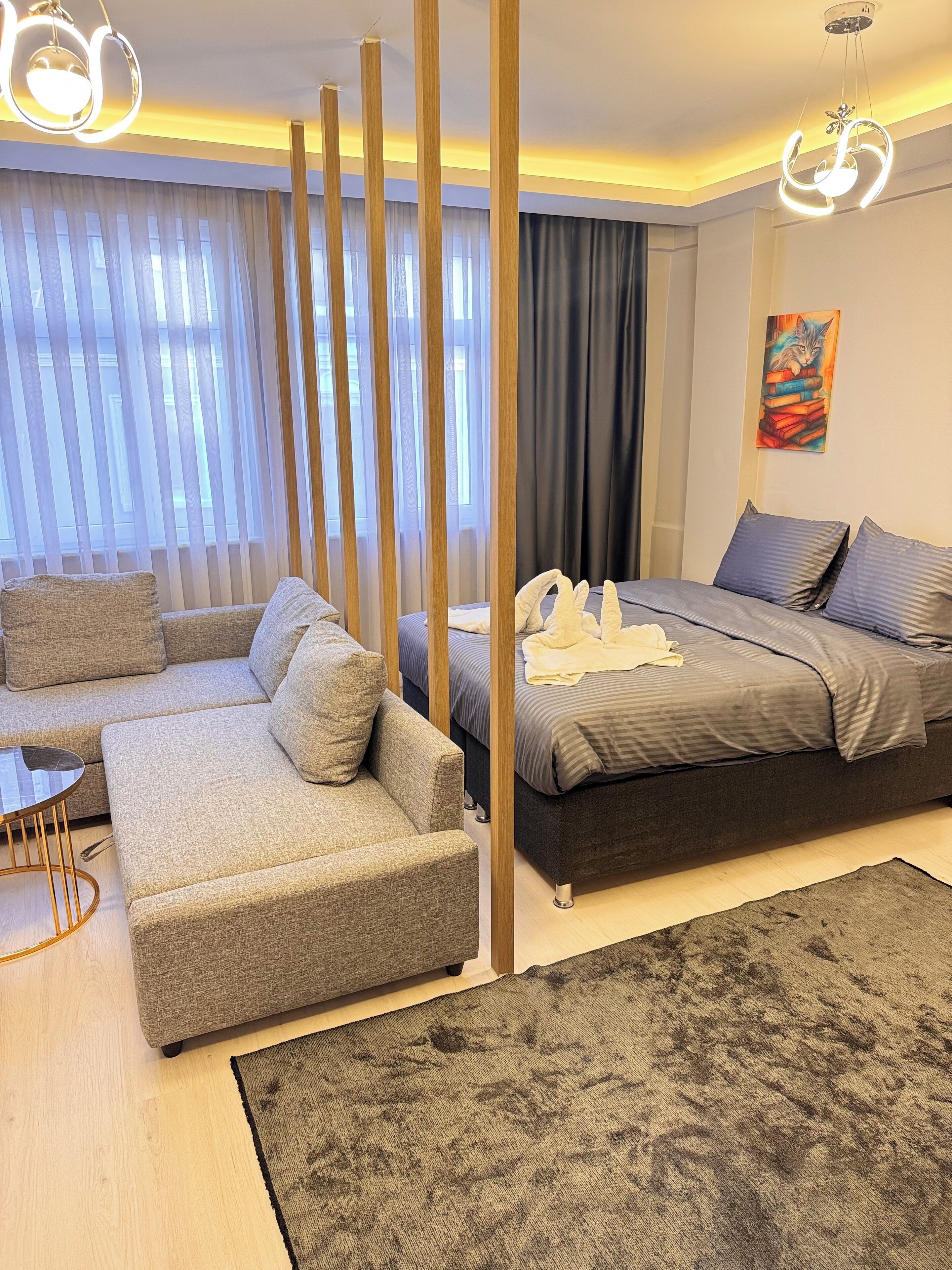 A modern bedroom with a grey sofa, a bed with grey bedding, a dark rug, and a painting of books on the wall.