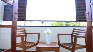 A balcony with two wooden chairs and a small table, a white vase with flowers, and a view of greenery through a window with shutters. - Seaport Negombo (Negombo)