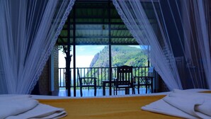 A room with a bed, white curtains, a balcony with chairs, and a view of a mountainous landscape.