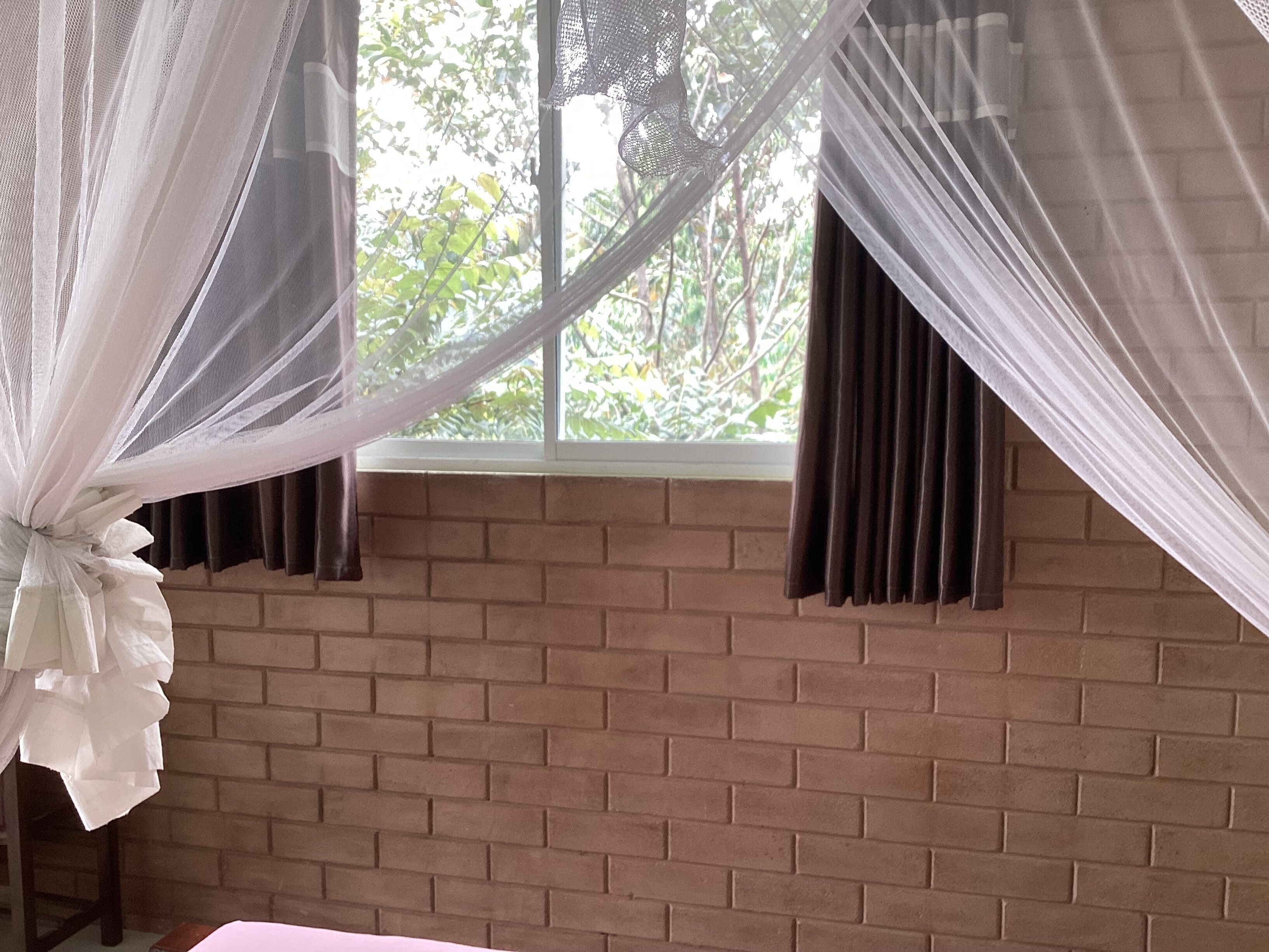 Una habitación con pared de ladrillo, ventana con cortinas y cama con cubrecolcha blanco.