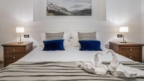 Uma cama bem arrumada com uma pintura de paisagem montanhosa acima, duas mesas de cabeceira com abajures, e um cobertor listrado sobre a cama.