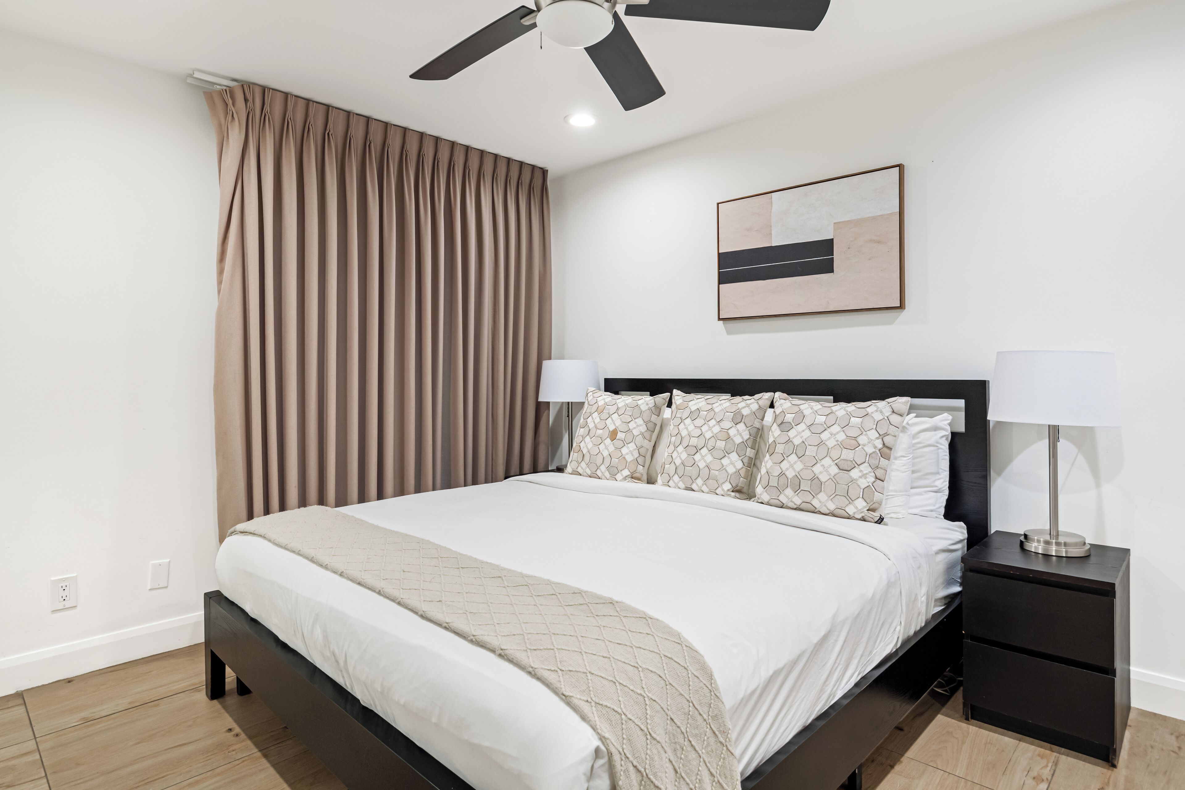 Une chambre à coucher moderne, avec un lit, des tables de chevet, un ventilateur de plafond et une œuvre d’art accrochée au mur.