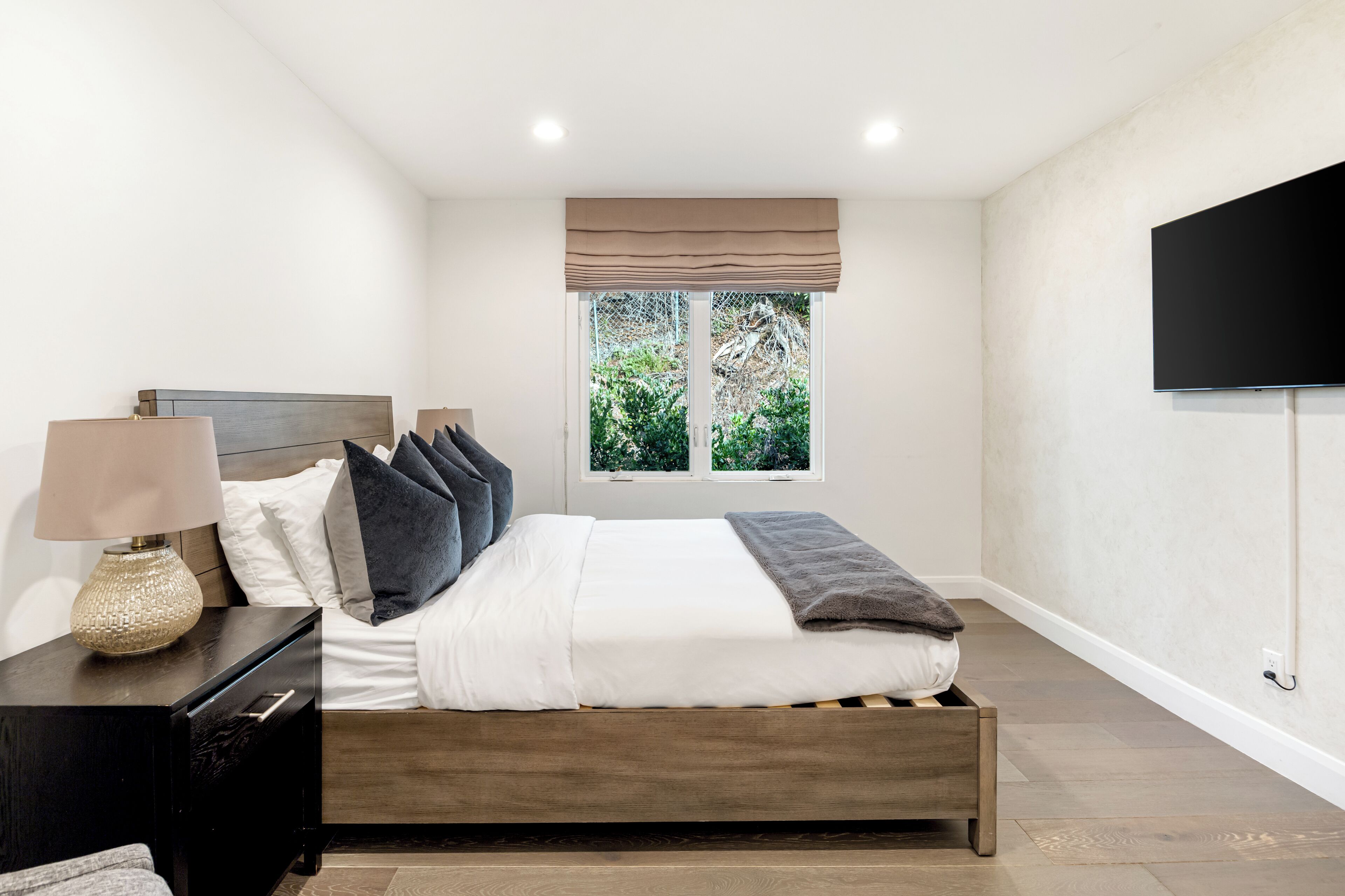 Une chambre à coucher comprenant un lit, une table de chevet, une lampe, une fenêtre donnant sur un espace verdoyant et un téléviseur fixé au mur.