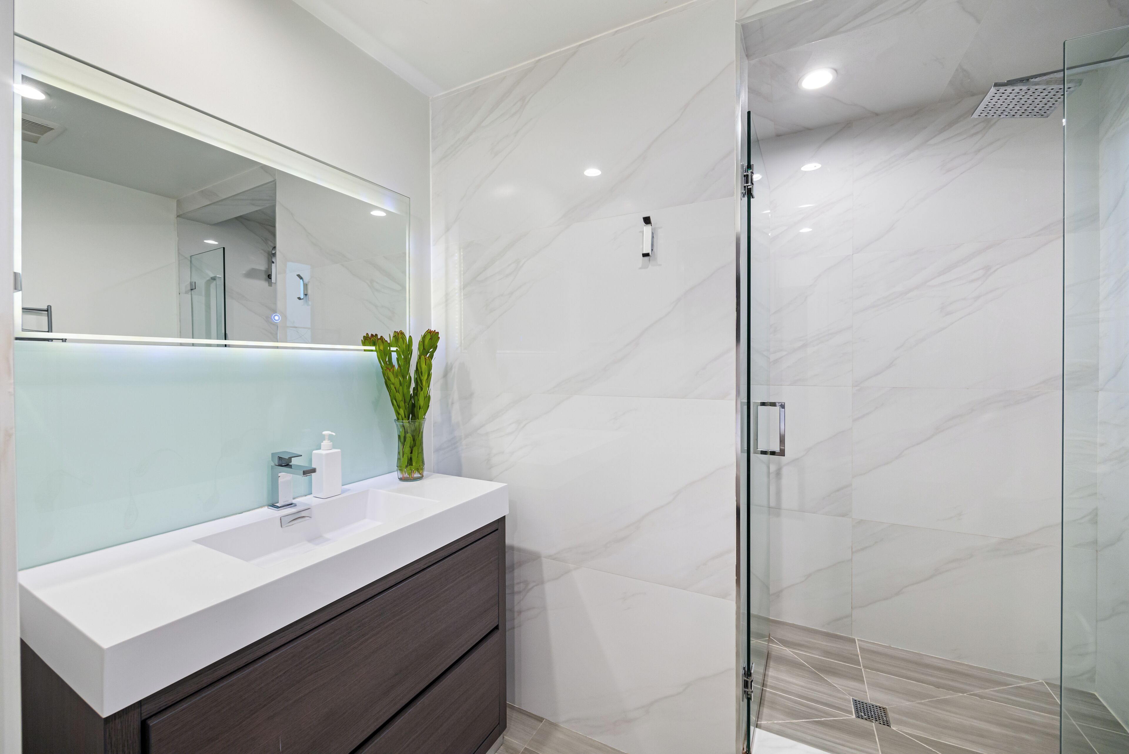 A modern bathroom with a white vanity, a glass shower enclosure, and marble walls.