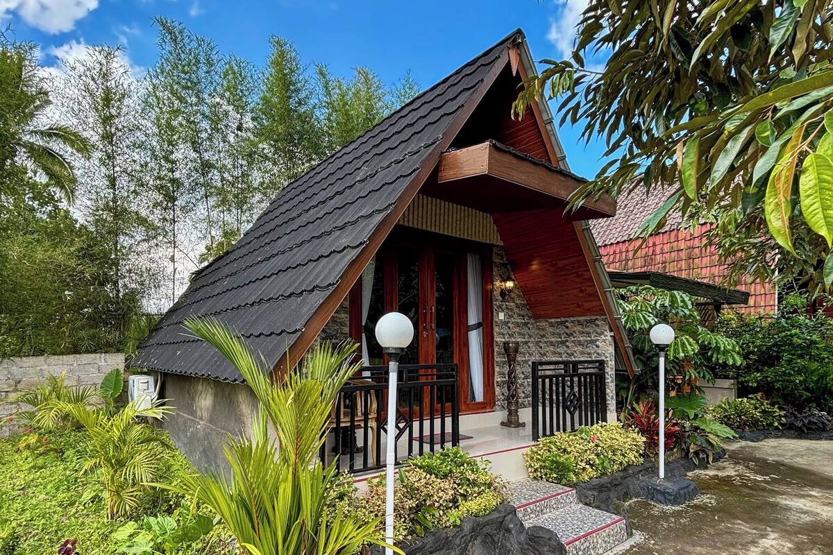 A small, rustic house with a sloped roof, surrounded by lush greenery and a stone wall.