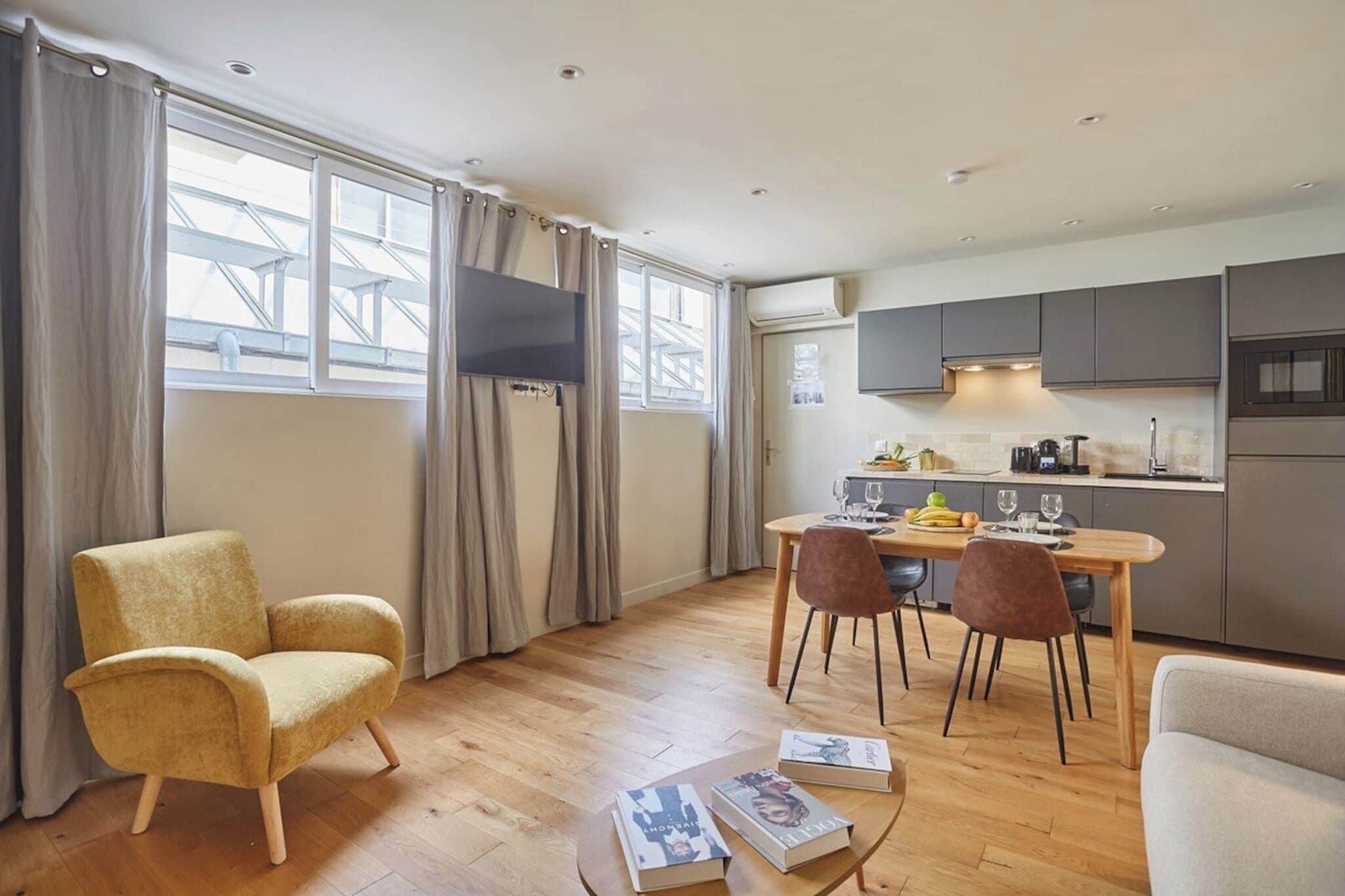 Un salon moderne comprenant un canapé, une table à manger avec des chaises, un téléviseur fixé au mur et un coin cuisine avec des placards et des appareils électroménagers.