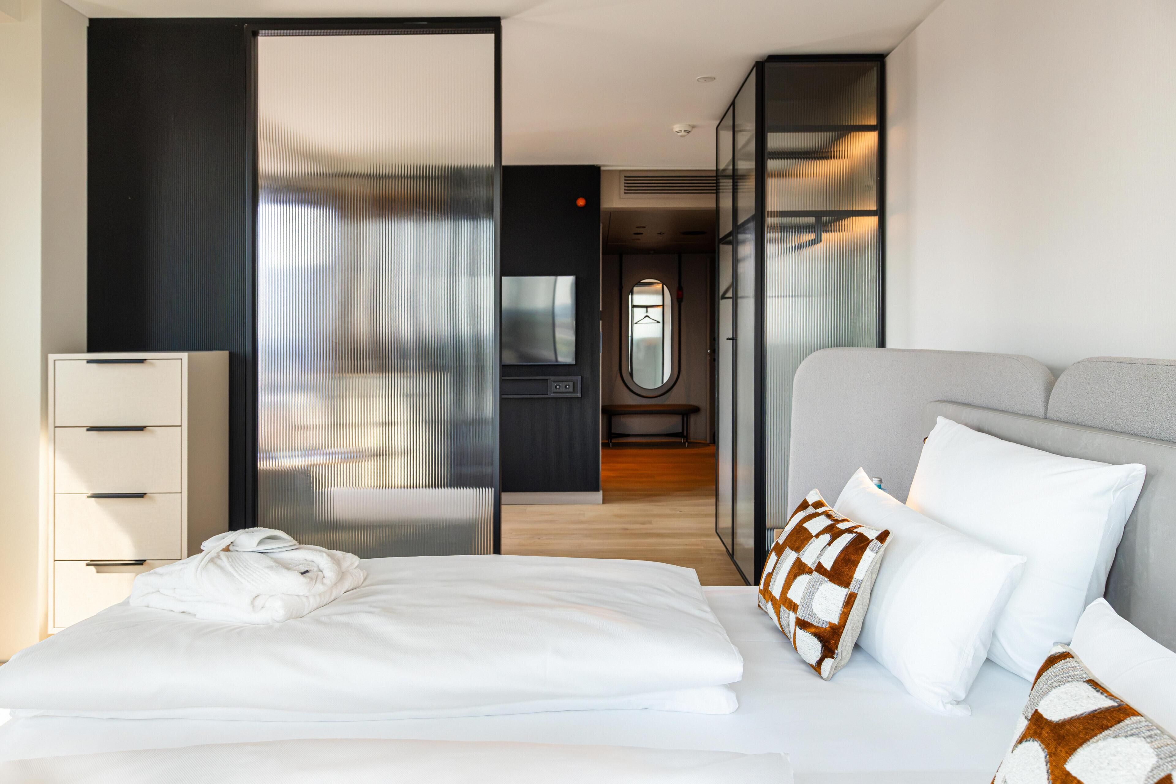 A modern hotel room with a bed, a bedside table, a wardrobe, and a bathroom visible through a glass partition.