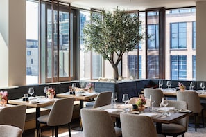 A restaurant with multiple tables, chairs, and a large tree in the center. - ATLANTIC Hotel Frankfurt - New opening (Frankfurt)