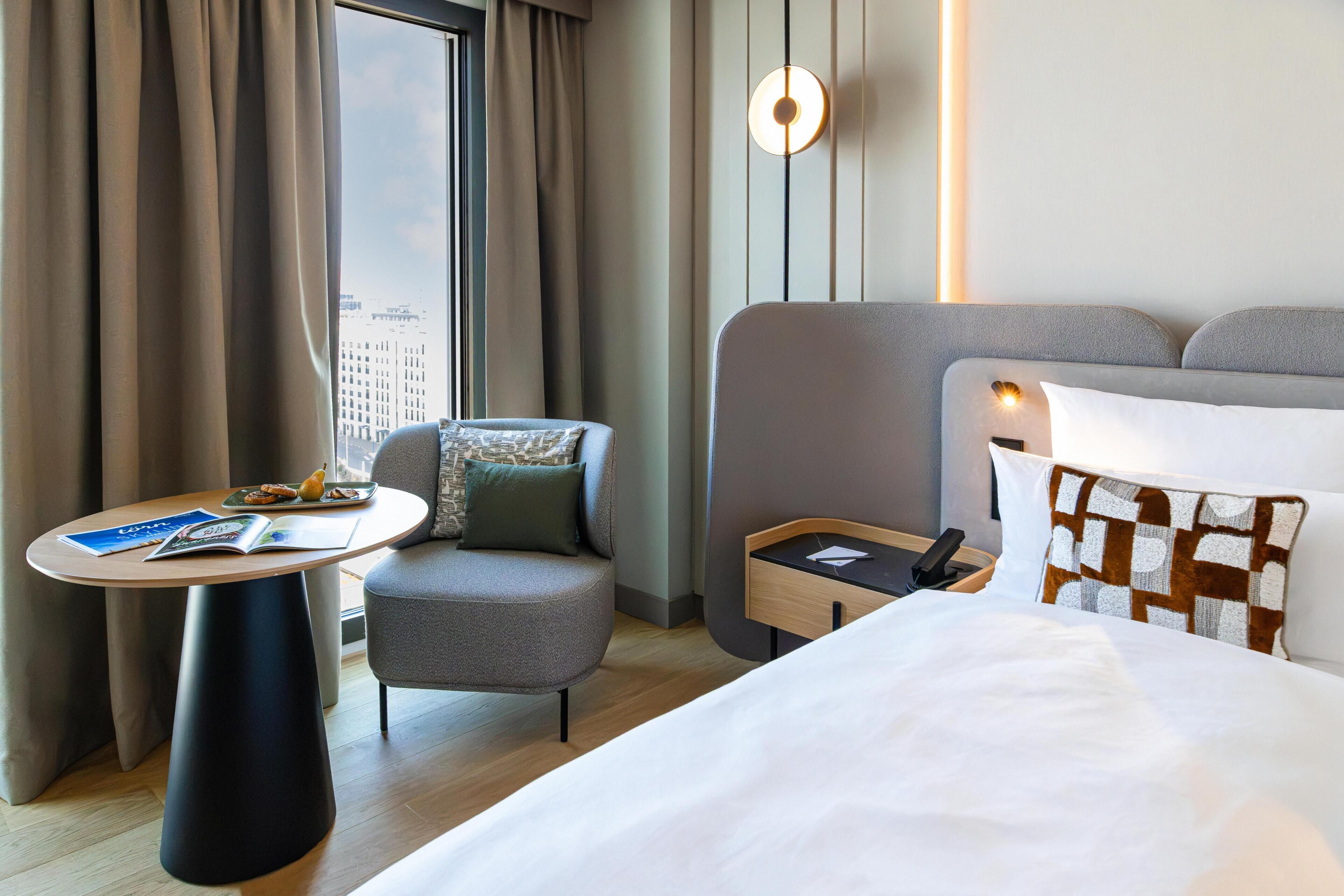 Een hotelkamer met een bed, een kleine tafel, een stoel, een bureau en een raam met gordijnen.