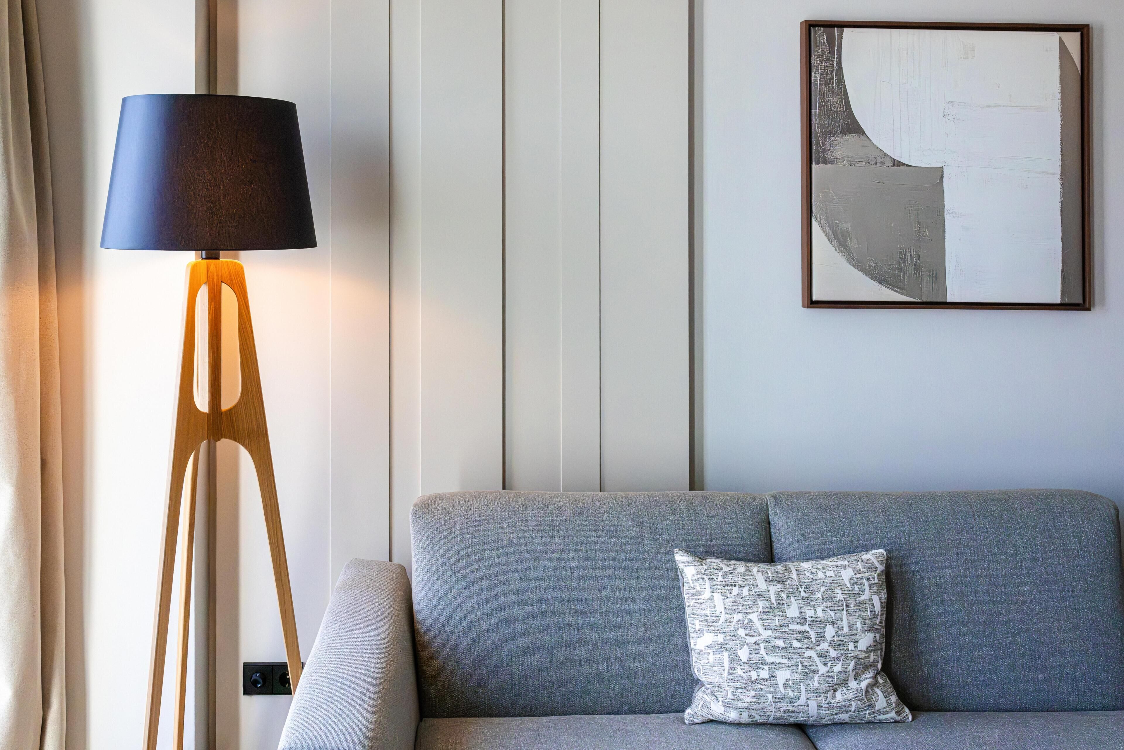 Una habitación con un sofá gris, un cojín estampado, una lámpara de pie con pantalla oscura y una obra de arte abstracta en la pared.