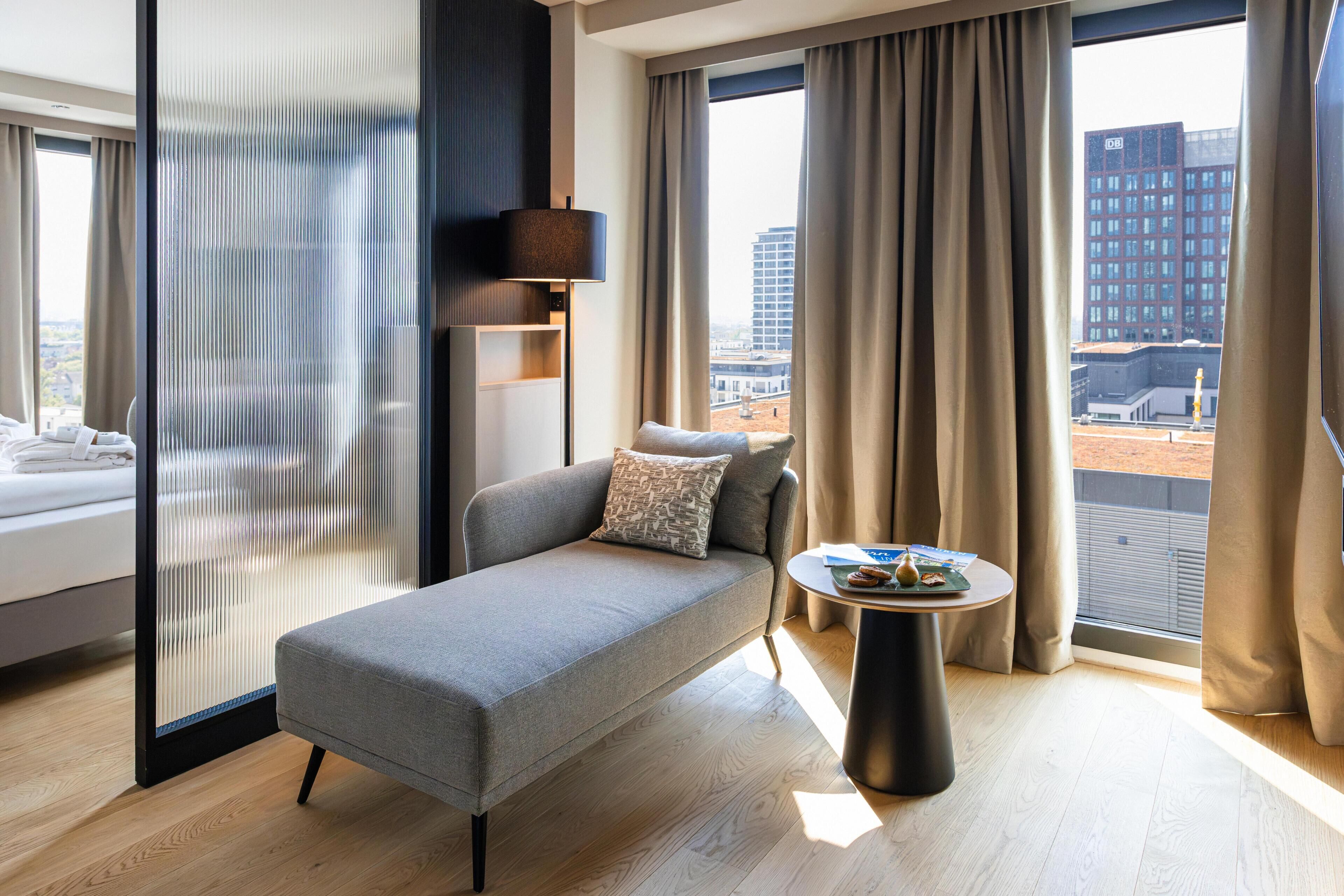 Ein modernes Hotelzimmer mit einem grauen Sofa, einem kleinen runden Tisch mit einem Buch und einer Obstplatte, einem großen Fenster mit Vorhängen und Blick auf die Stadt.