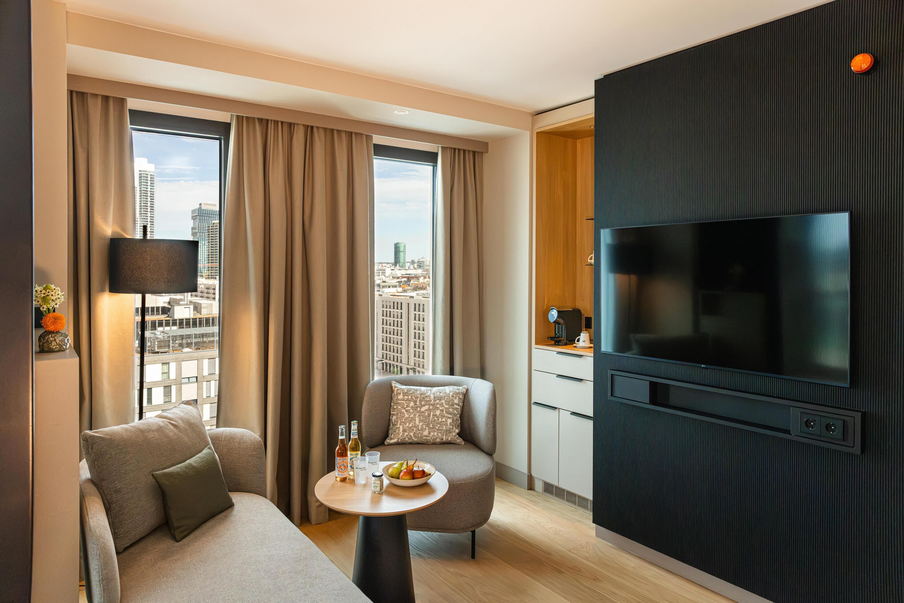 A modern hotel room with a sofa, a small table with a bowl of fruit, a coffee machine, and a TV mounted on a dark panel.