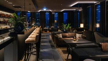A modern bar area with a long counter, bar stools, and a city view through large windows.