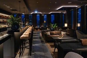 Zona de bar moderna con una barra alargada, taburetes y vistas a la ciudad a través de amplios ventanales.