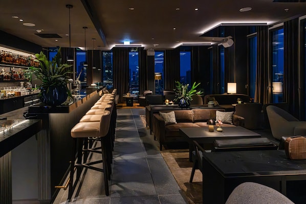 A modern bar area with a long counter, bar stools, and a city view through large windows. - ATLANTIC Hotel Frankfurt - New opening (Frankfurt)