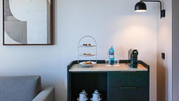 Habitación de hotel con kitchenette, cafetera, botella de agua y un cuadro abstracto enmarcado.