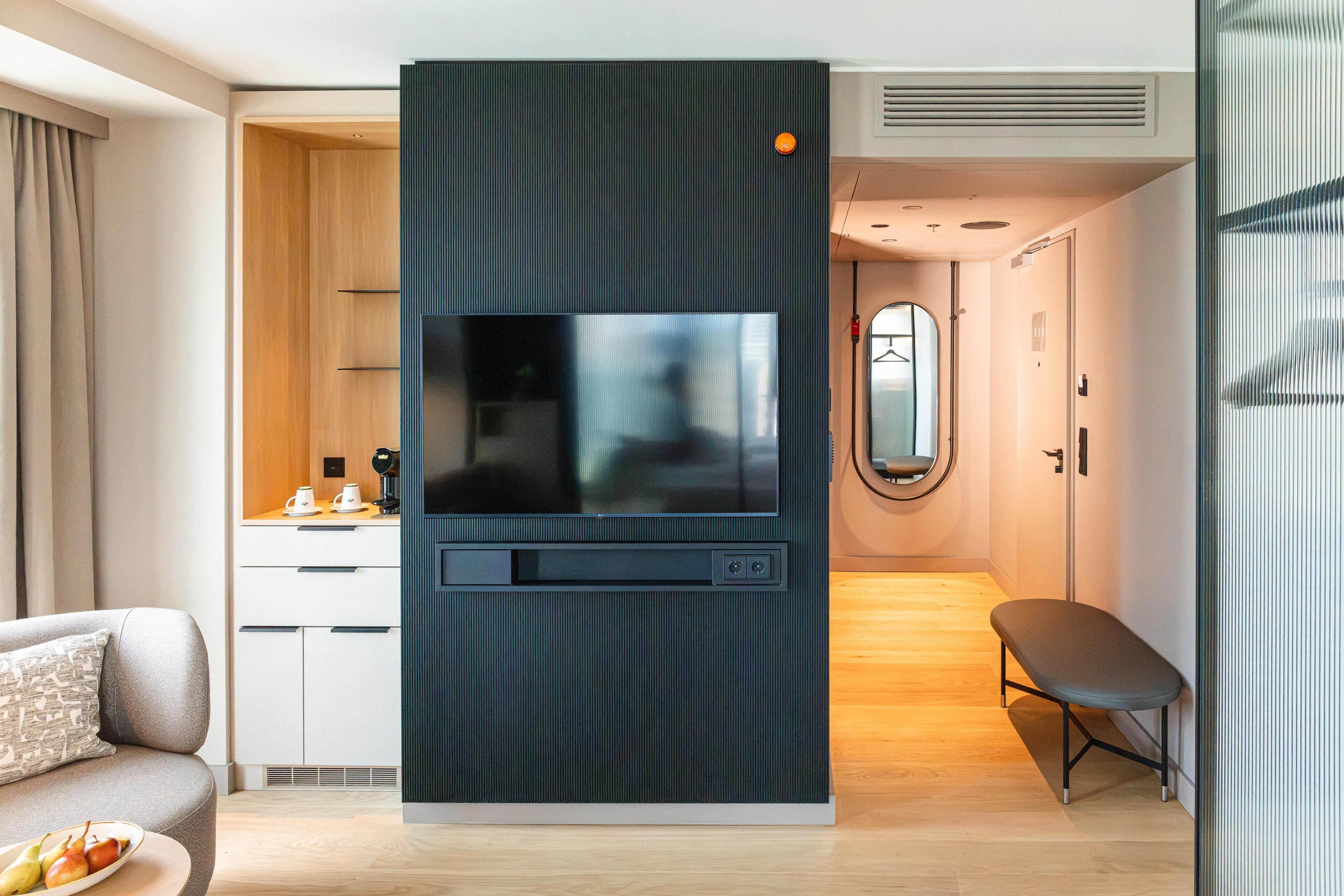 A modern hotel room with a flat-screen TV mounted on a dark panel, a wooden desk with a coffee maker, a sofa with a cushion, and a round mirror on the wall.