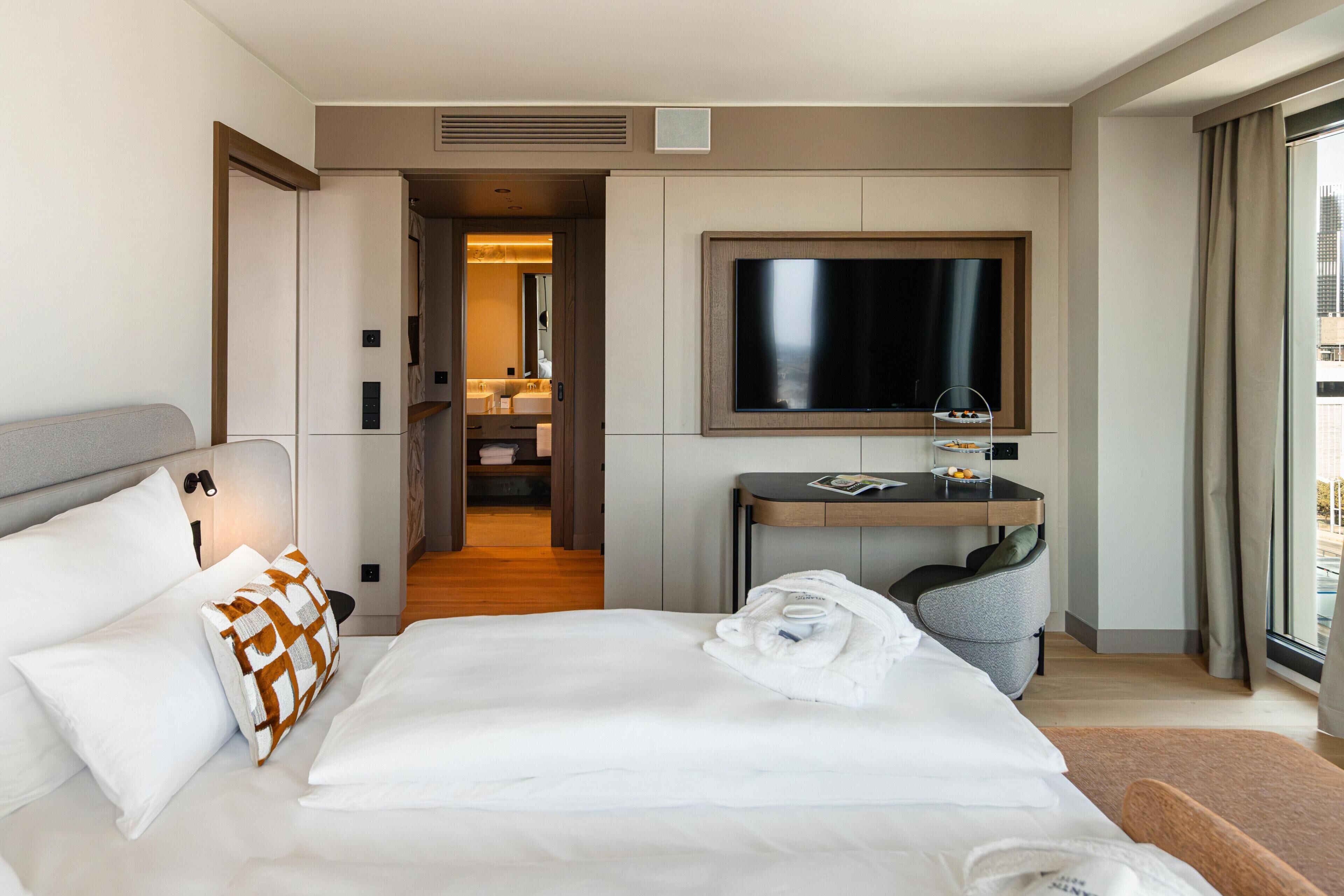 A hotel room with a large bed, a desk with a flat-screen TV, a bathroom with a mirror, and a view of the city through the window.