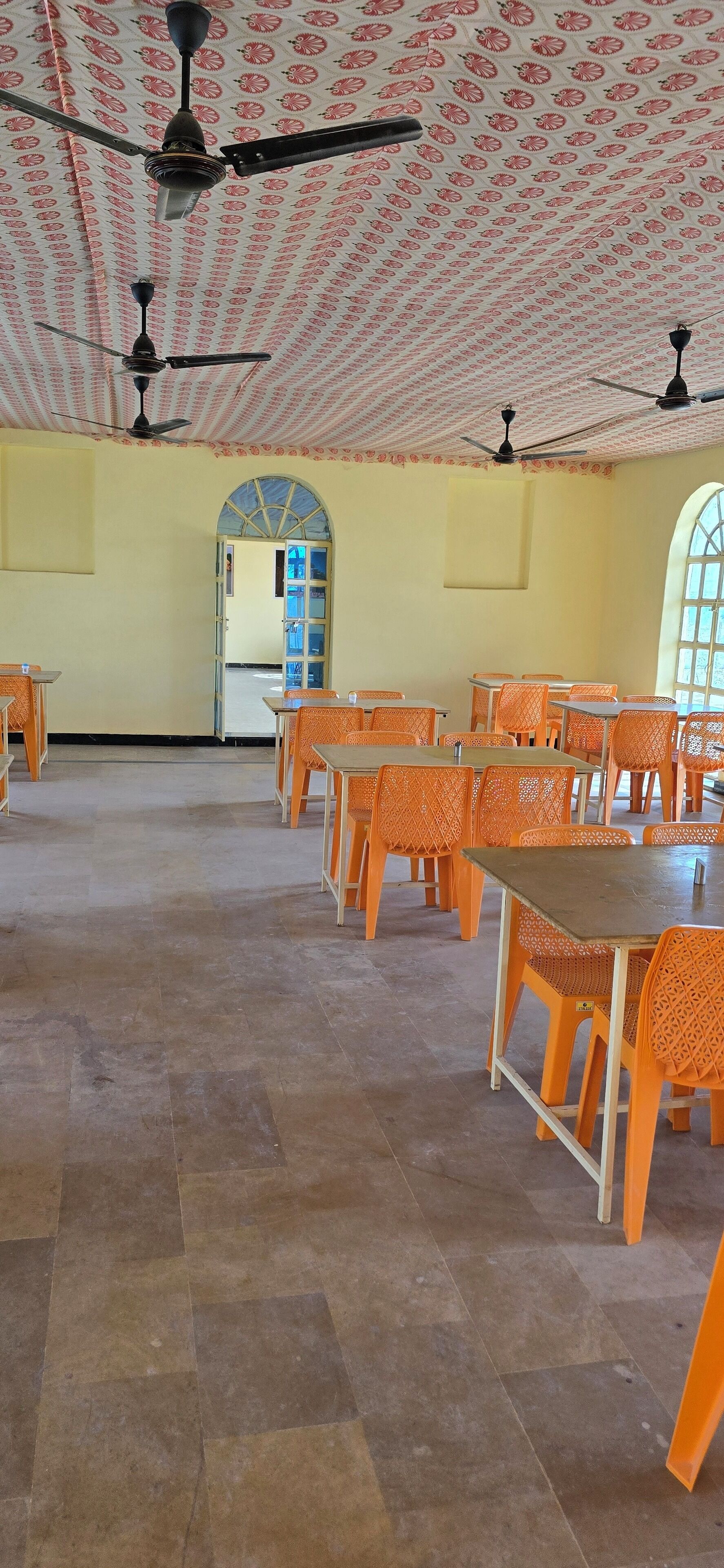 A spacious hall with orange chairs and tables, a patterned ceiling, and a large arched doorway.