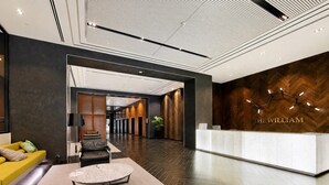 A modern hotel lobby with a reception desk, a seating area with a sofa and armchair, and a hallway leading to elevators. - Super King Beds, Vinyl + Pool | CBD Luxury (Melbourne)