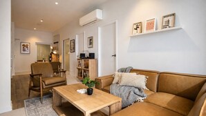 A modern living room with a sofa, armchair, and coffee table. There is a bookshelf and a wall-mounted air conditioner. - Super King Beds, Vinyl + Pool | CBD Luxury (Melbourne)