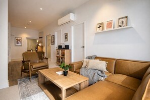 A modern living room with a sofa, armchair, and coffee table. There is a bookshelf and a wall-mounted air conditioner. - Super King Beds, Vinyl + Pool | CBD Luxury (Melbourne)