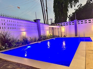 Une piscine rectangulaire à l’eau bleutée, entourée d’une clôture blanche percée de fentes verticales, et un chemin de pierre menant à la piscine.