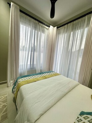 Een slaapkamer met een groot bed, lichte gordijnen en een plafondventilator.