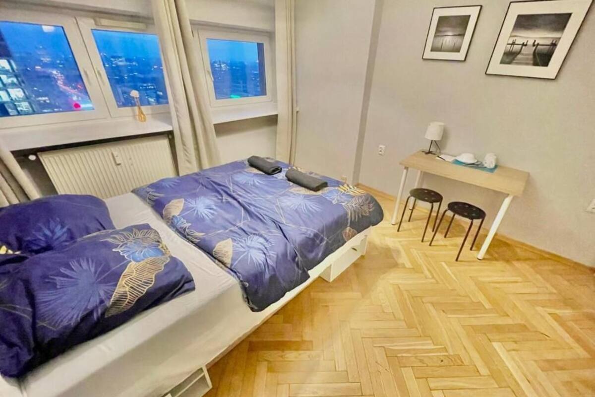 A bedroom with a bed, a desk, and a view of the city through windows.