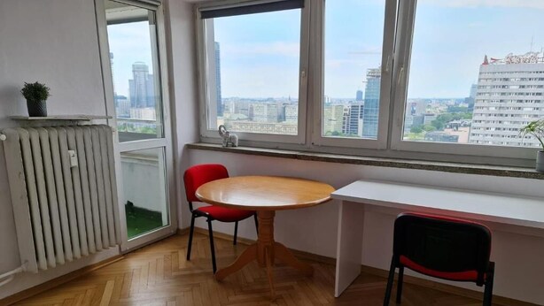 A room with a wooden floor, a white desk, a red chair, and a view of the city through large windows. - Fantastic Inn Warsaw - CH35 (Warsaw)