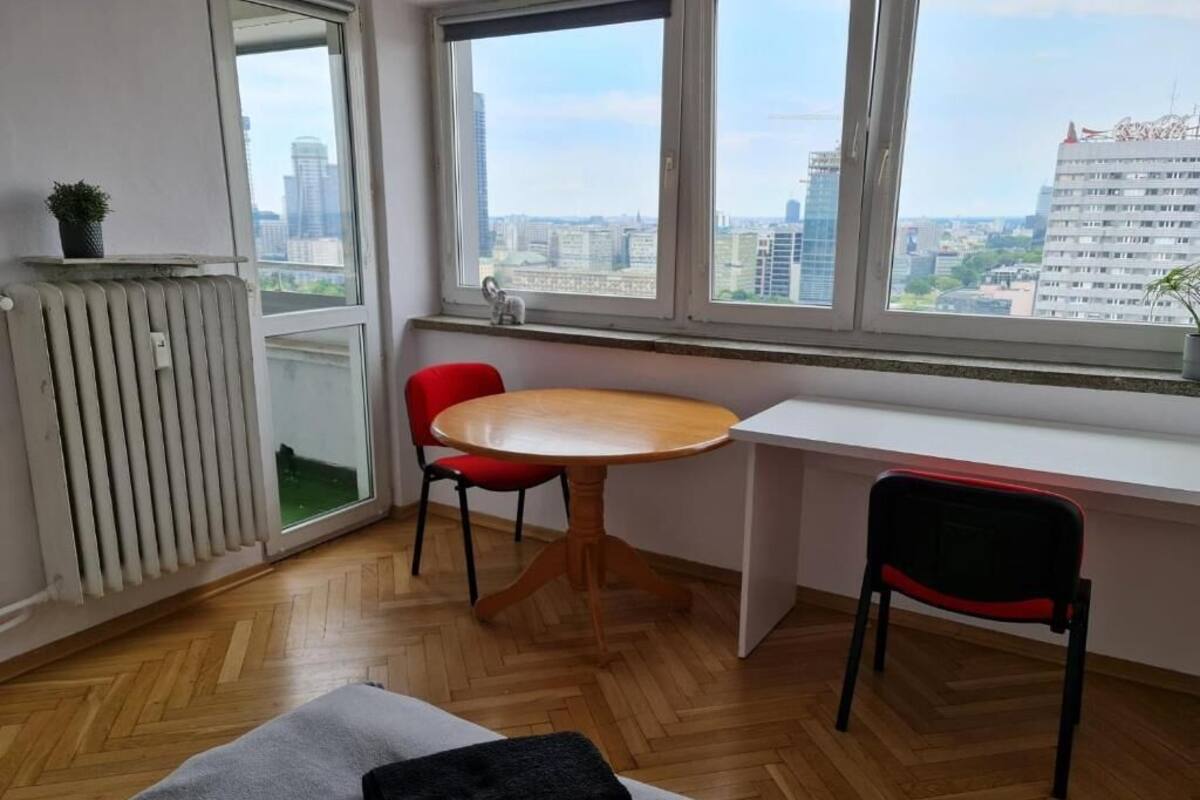 A room with a wooden floor, a white desk, a red chair, and a view of the city through large windows.