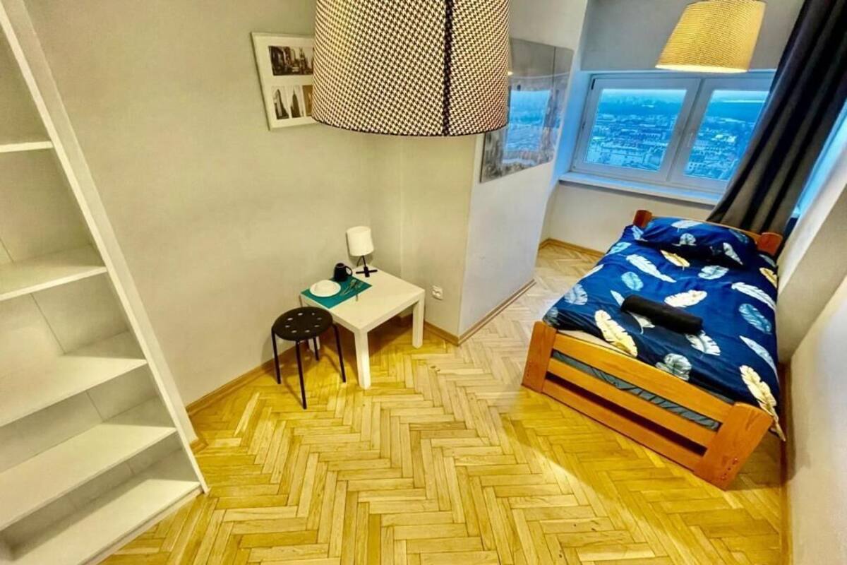 A bedroom with a herringbone floor, a bed with a blue cover, a small table with a lamp, and a window with a city view.