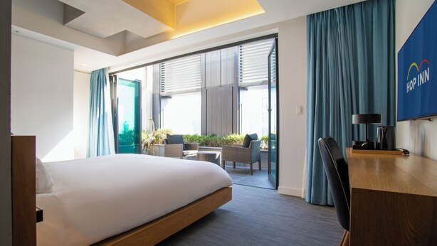 A modern hotel room with a bed, a desk, a chair, and a view of a balcony with plants. - HOP INN Seoul Myeongdong (Seoul)