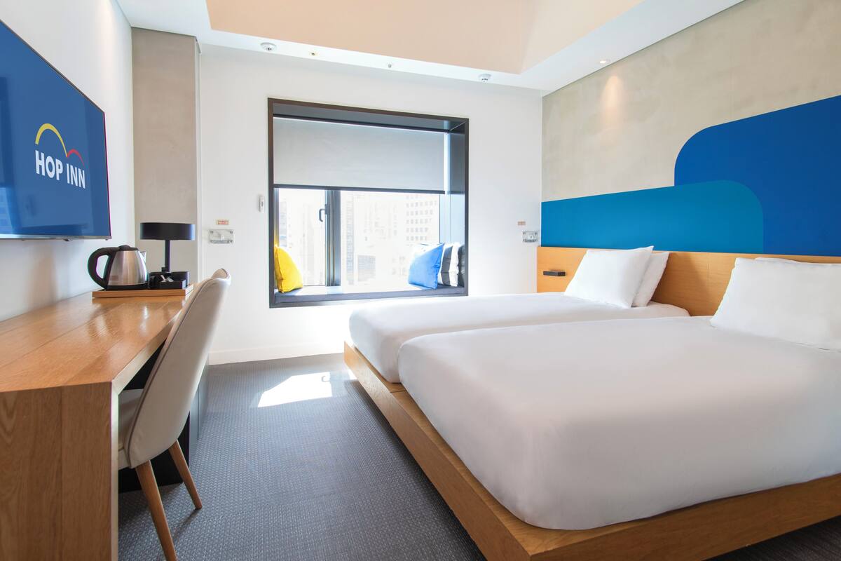 Ein Hotelzimmer mit zwei Betten, einem Schreibtisch, einem Sessel, einem Fernseher und einem Fenster mit Blick auf die Stadt.
