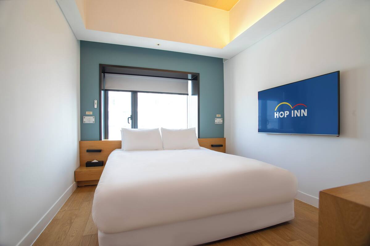 Ein modernes Hotelzimmer mit einem großen Bett, einem Fenster mit Jalousien, einem Holznachttisch und einem an der Wand befestigten Schild für Hop Inn.