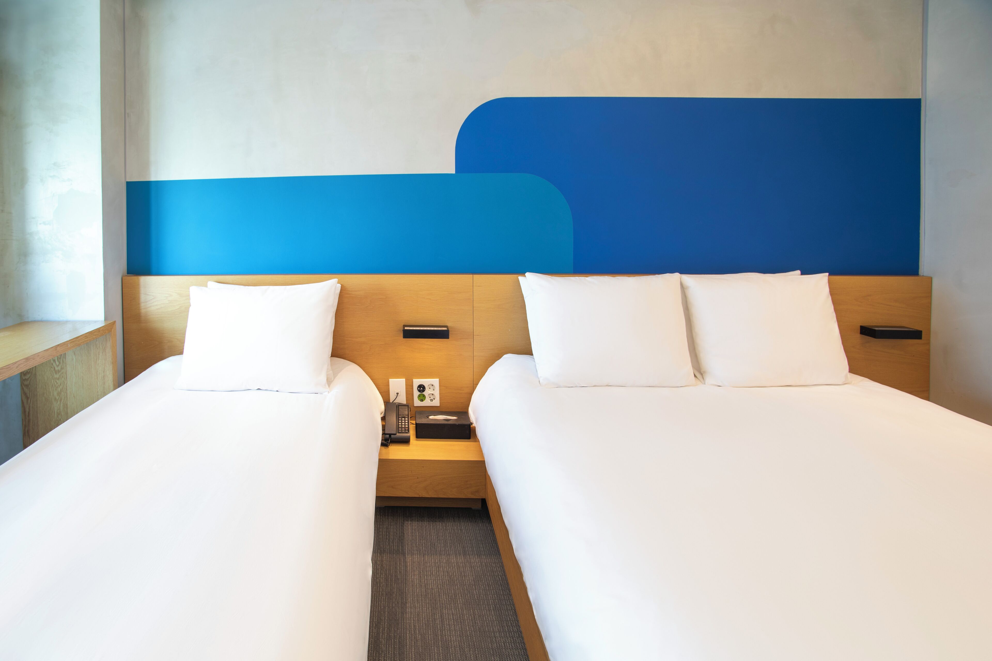 Habitación de hotel con dos camas, cabecera de madera, mesita de noche con teléfono y pañuelos, y una pared decorativa azul.
