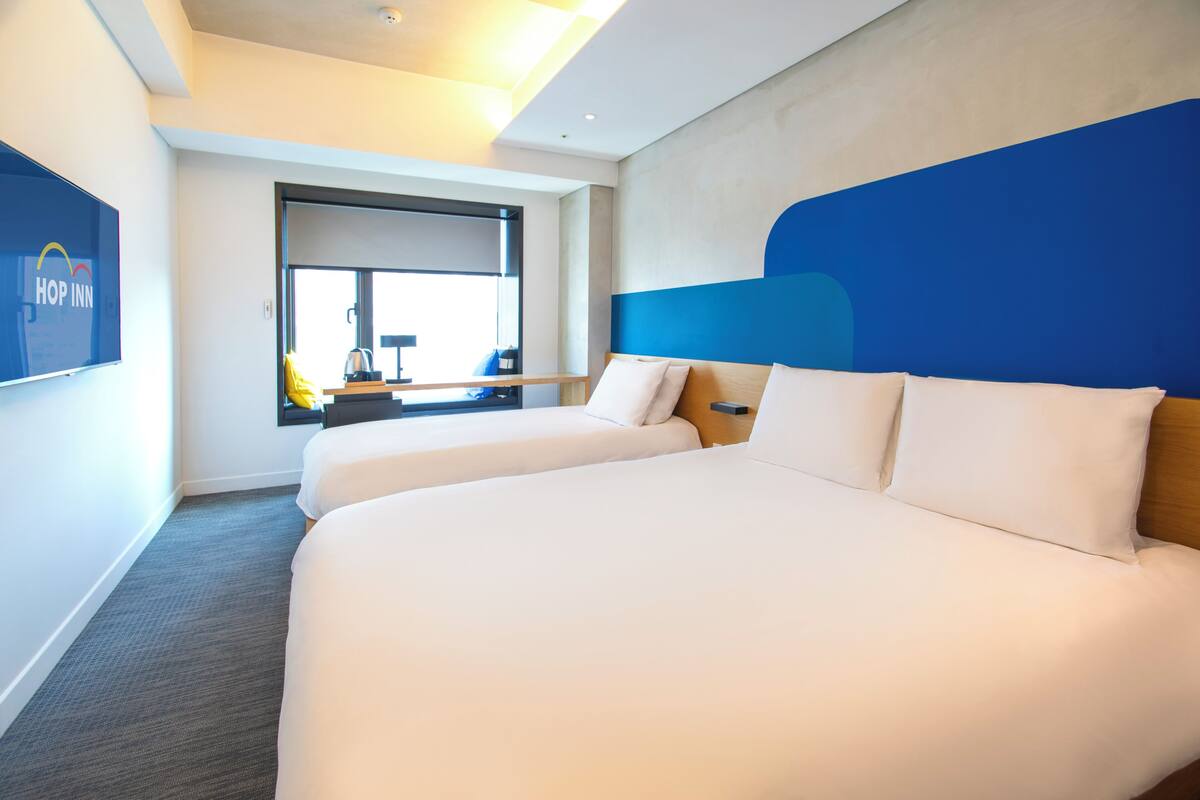 Ein Hotelzimmer mit zwei Betten, einem großen Fenster, einem Schreibtisch mit Stuhl und einem an der Wand befestigten Bildschirm.