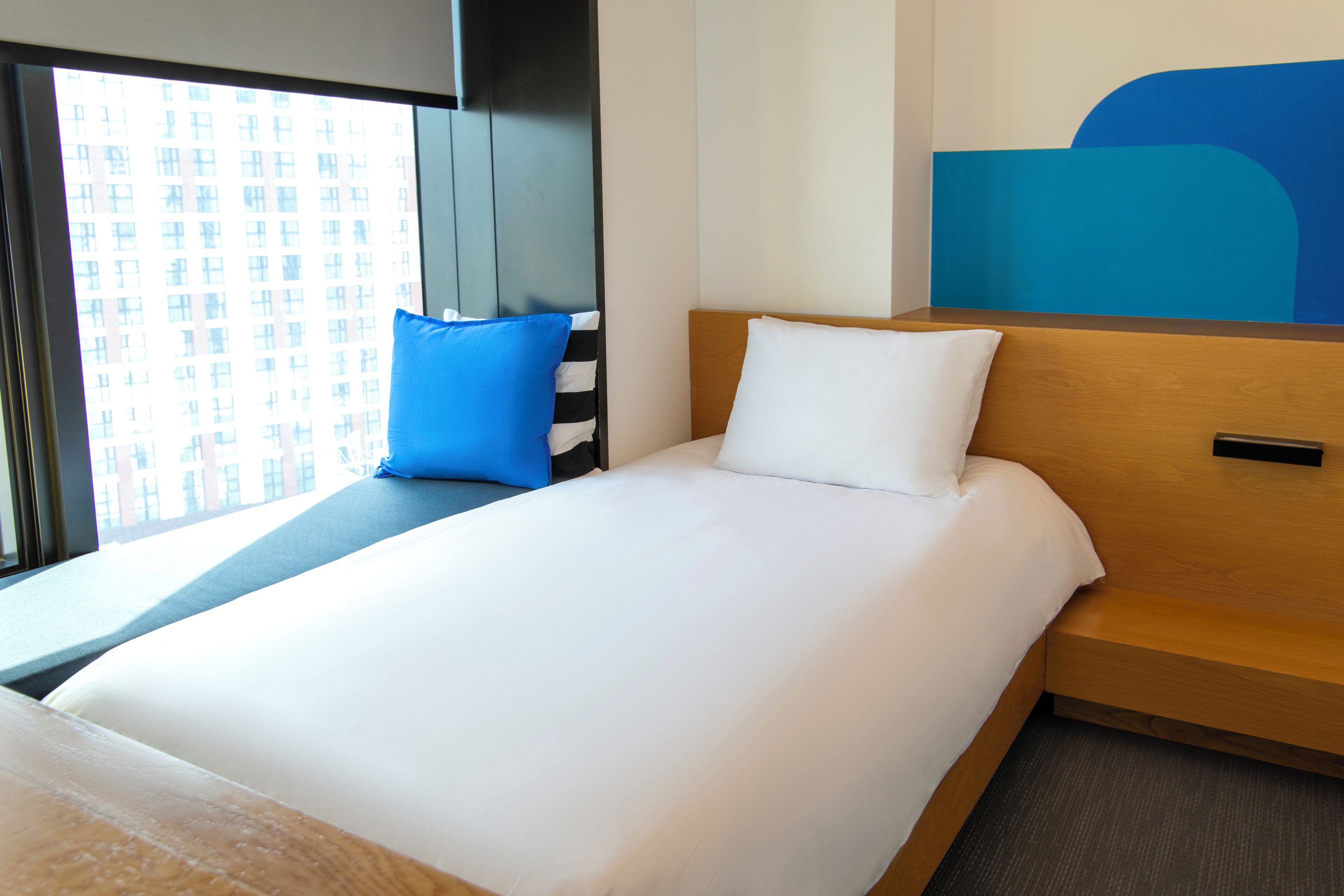 Une chambre d’hôtel avec un lit, une fenêtre, une vue sur les bâtiments et un mur d’accent bleu.