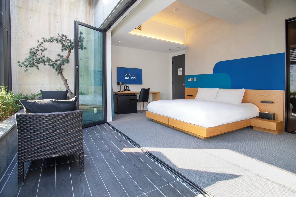Ein modernes Hotelzimmer mit einem großen Bett, einer Sitzecke mit einem Sessel, einem Schreibtisch und einer Glastür zum Außenbereich.