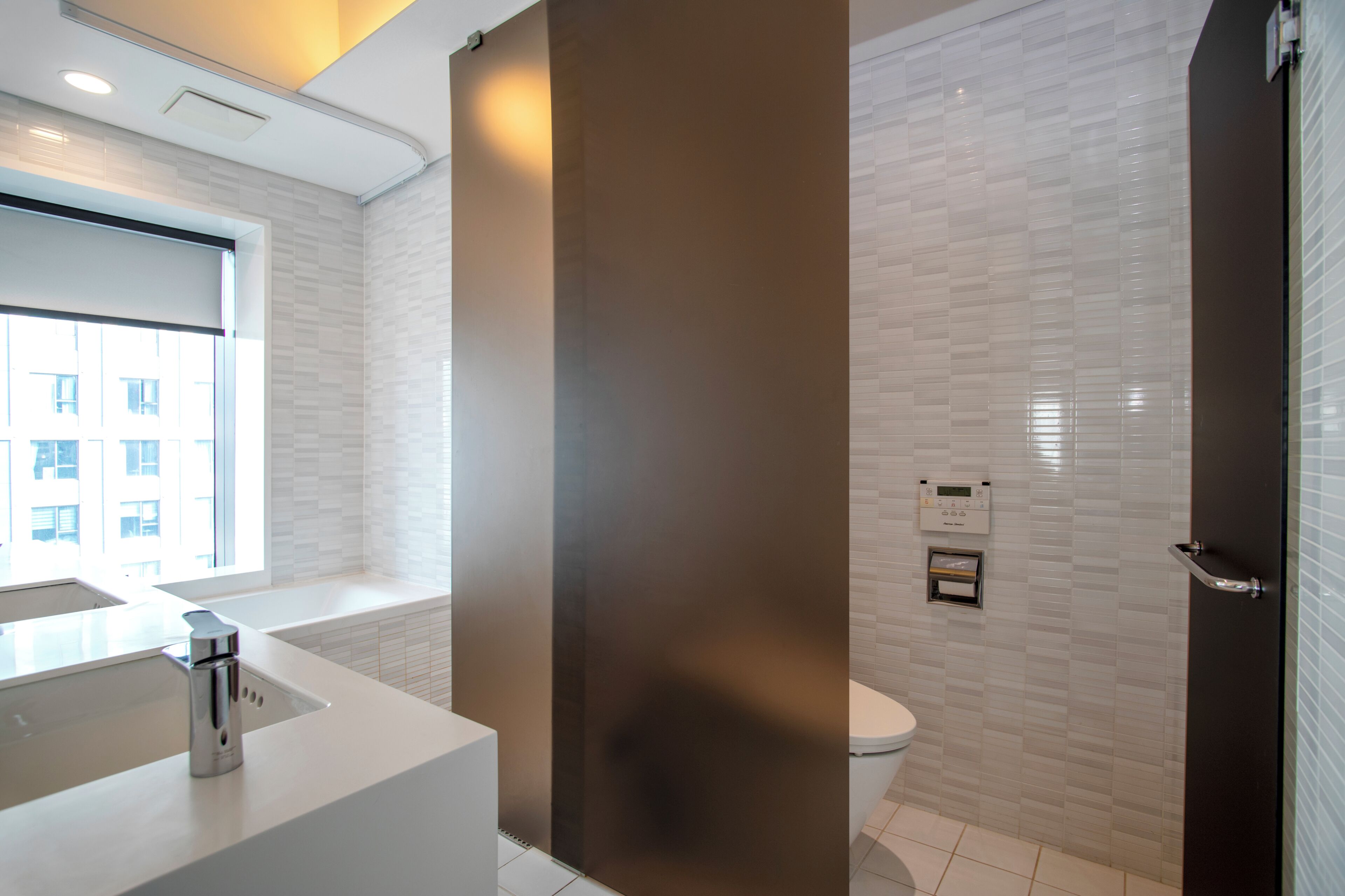 A modern bathroom with a white sink, toilet, and shower area, featuring a large window and a control panel on the wall.