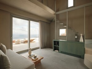 Een moderne woonkamer met een grote schuifpui naar een balkon met uitzicht op zee. Er staat een groene console tafel met decoratieve objecten en een boekenkast.