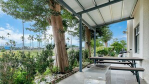 A covered patio area with a grill and table, surrounded by greenery and palm trees, overlooking a scenic landscape. - Longtian (Luye)
