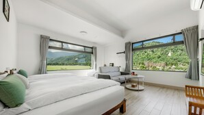 A modern bedroom with a large bed, a sofa, and a small table. There are large windows offering a view of a green landscape. - Longtian (Luye)