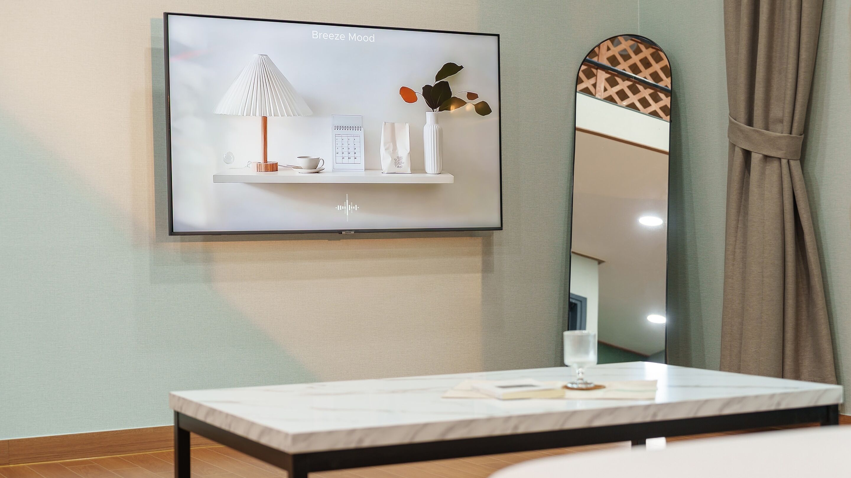 A modern hotel room with a flat-screen TV mounted on the wall displaying a 'Breeze Mood' scene, a small table with a book, and a mirror reflecting the room's decor.