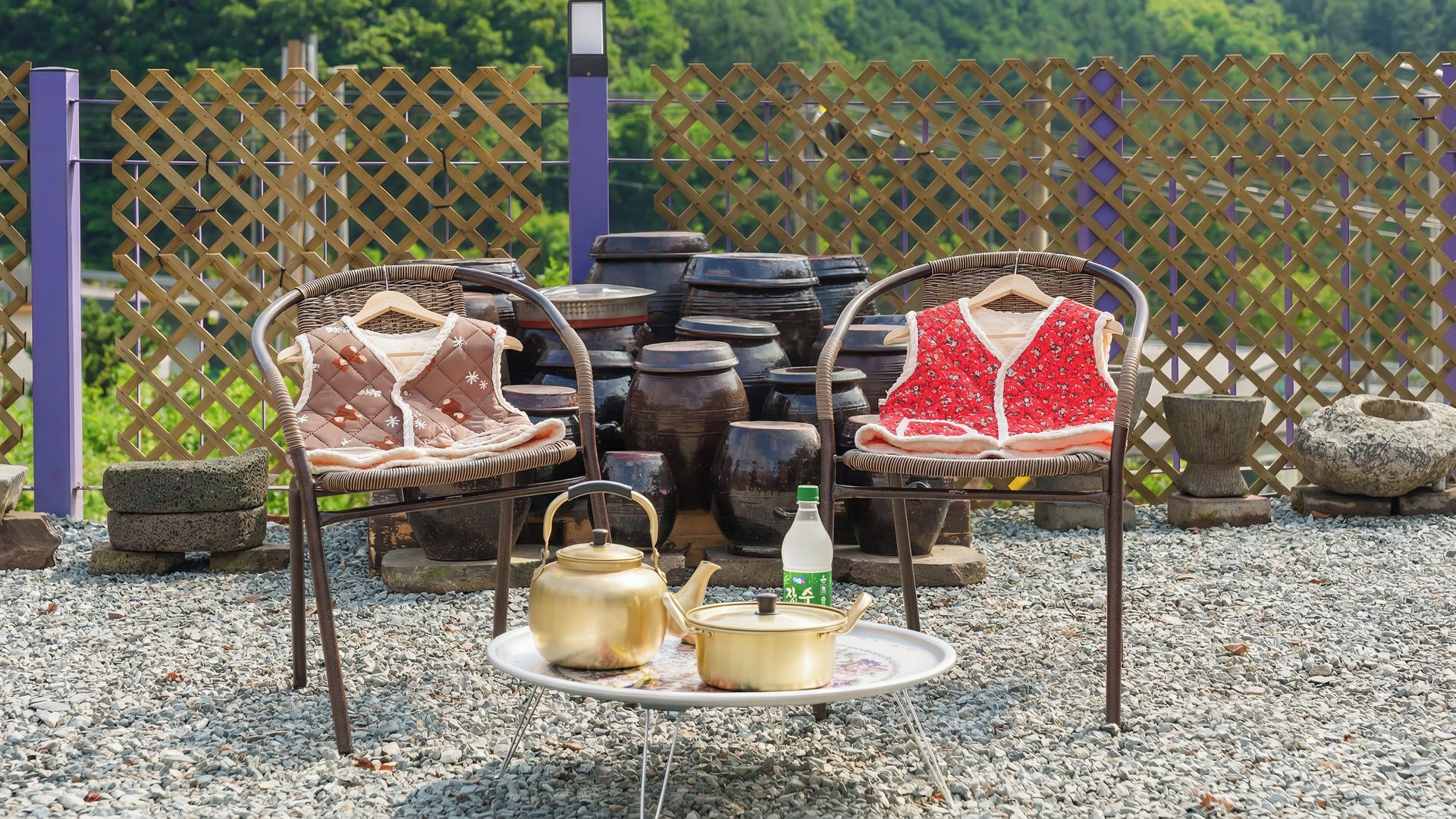 Zwei Korbstühle mit Decken, ein Metalltisch mit Teekanne und Flasche, sowie im Hintergrund Keramik und ein Zaun.