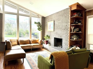 A living room with a brick fireplace, a sofa, a green armchair, and a bookshelf. - Midcentury Gem with Beautiful Outdoor Kitchen (Wheat Ridge)