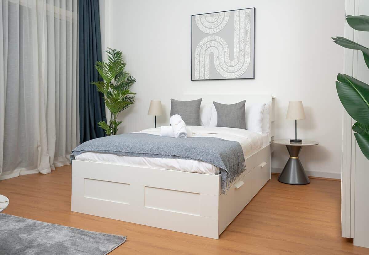 A modern bedroom with a white bed, grey bedding, a grey throw, two bedside lamps, a grey abstract wall art, and a potted plant.